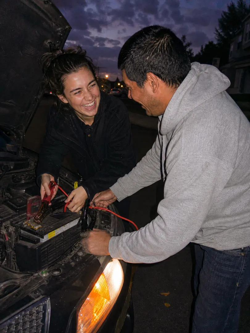 Two neighbors laughing while jump-starting a car at dusk